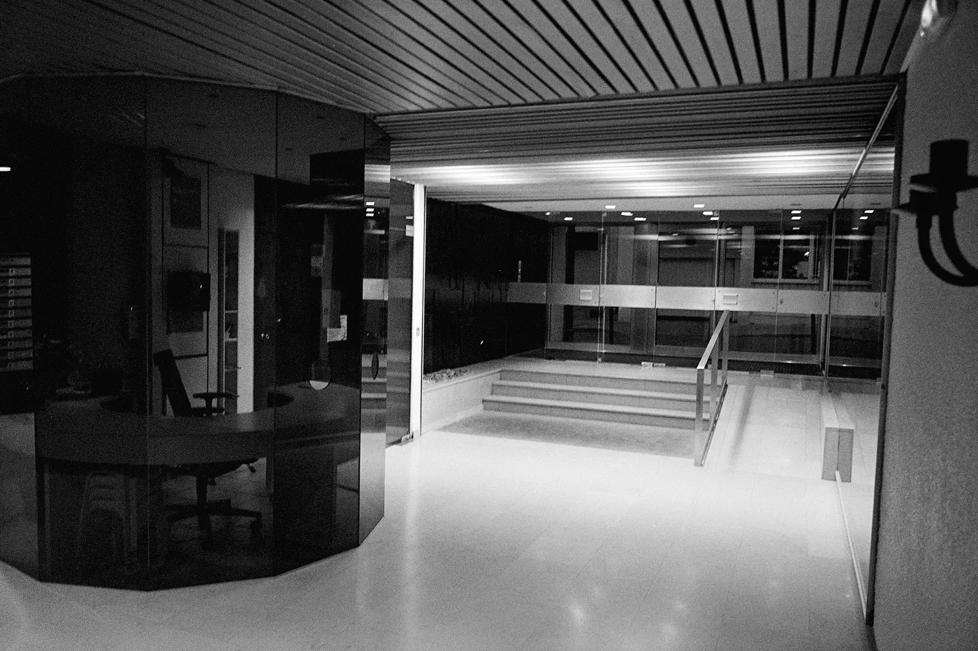 An empty office lobby in black and white: glass walls, a reception desk, steps up to a corridor.