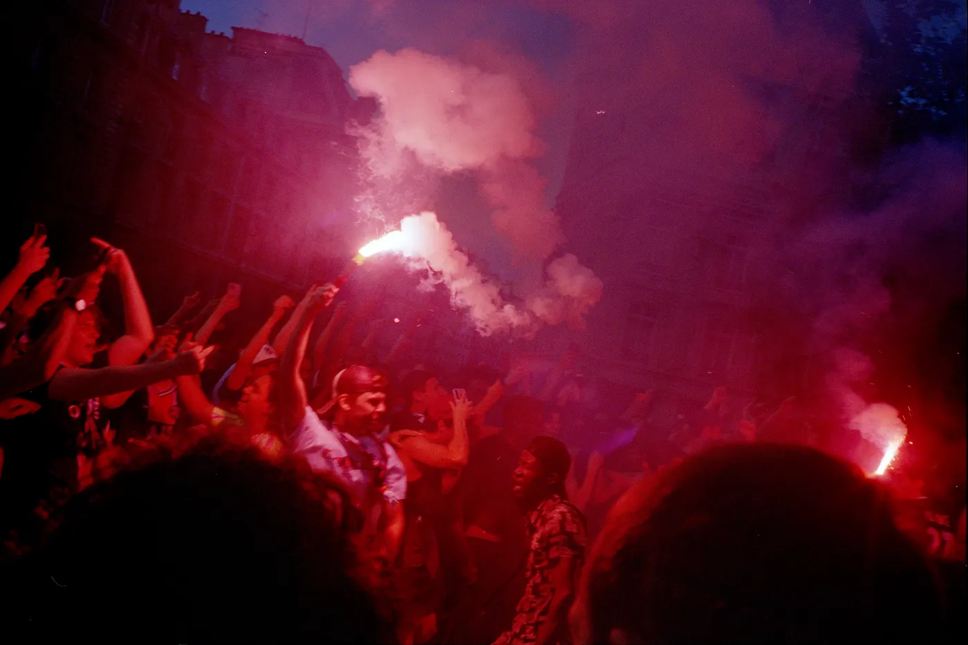 A night crowd under red smoke, arms raised, a flare burning above them.