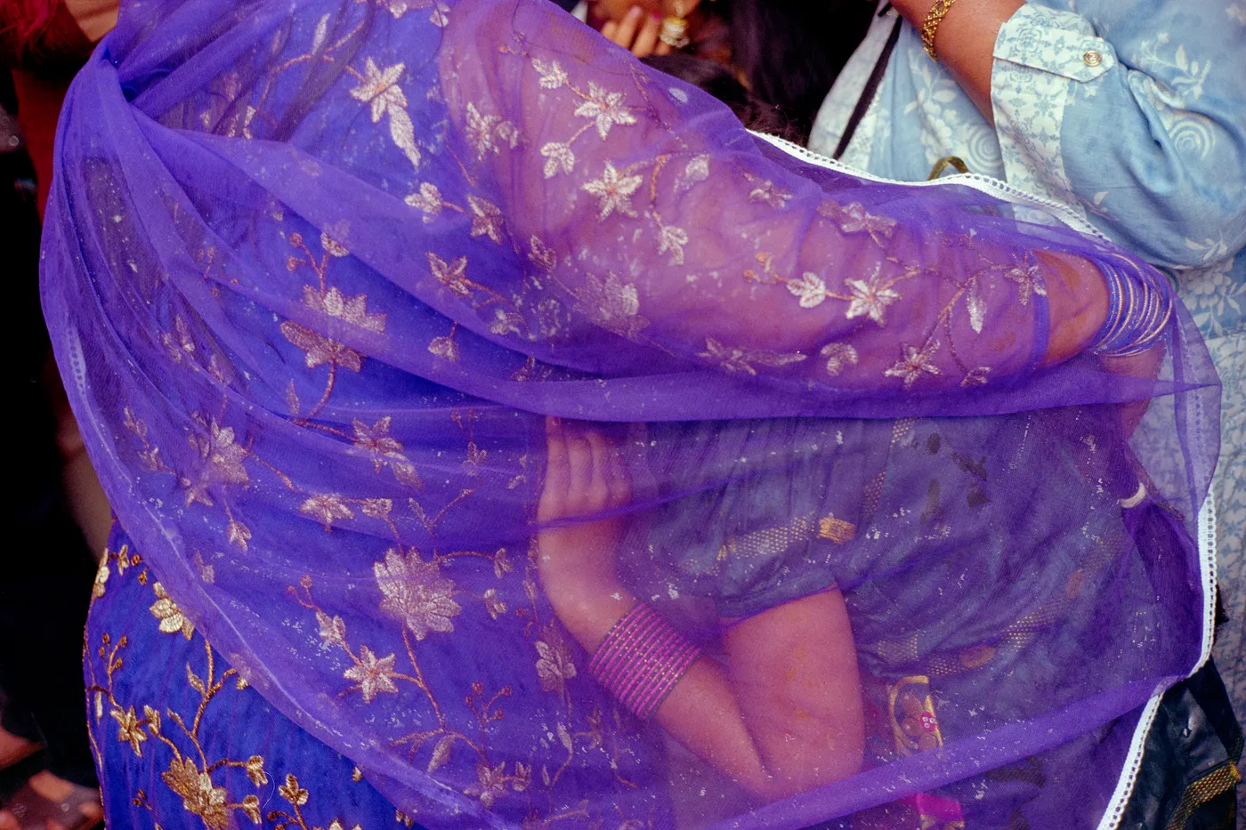 A purple embroidered sari veiling a face and a raised hand, in a night crowd.