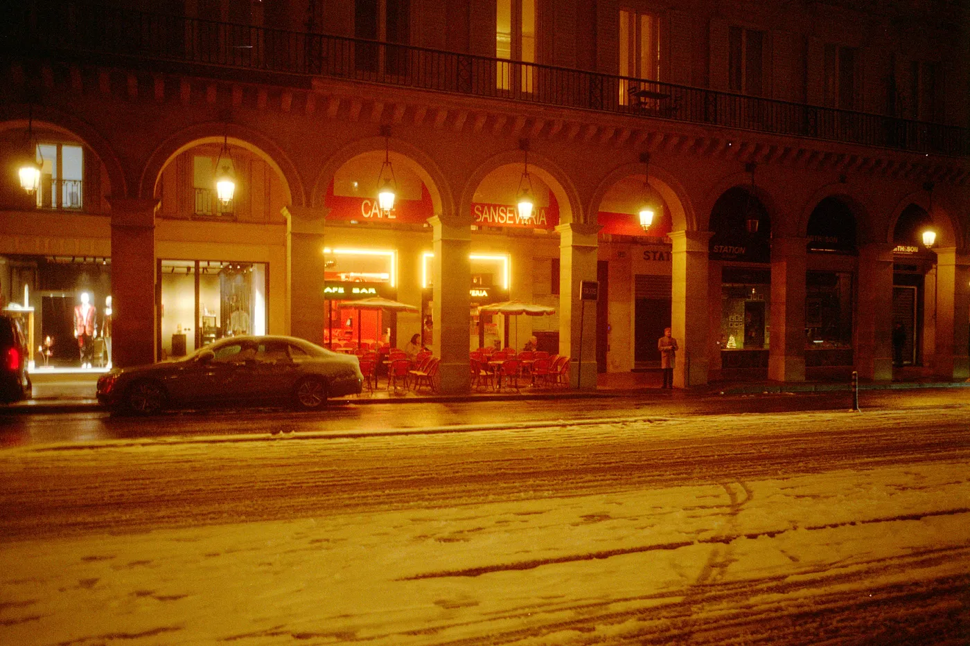 A snowy Paris street at night, a lit cafe under arcades, tyre tracks in the snow.