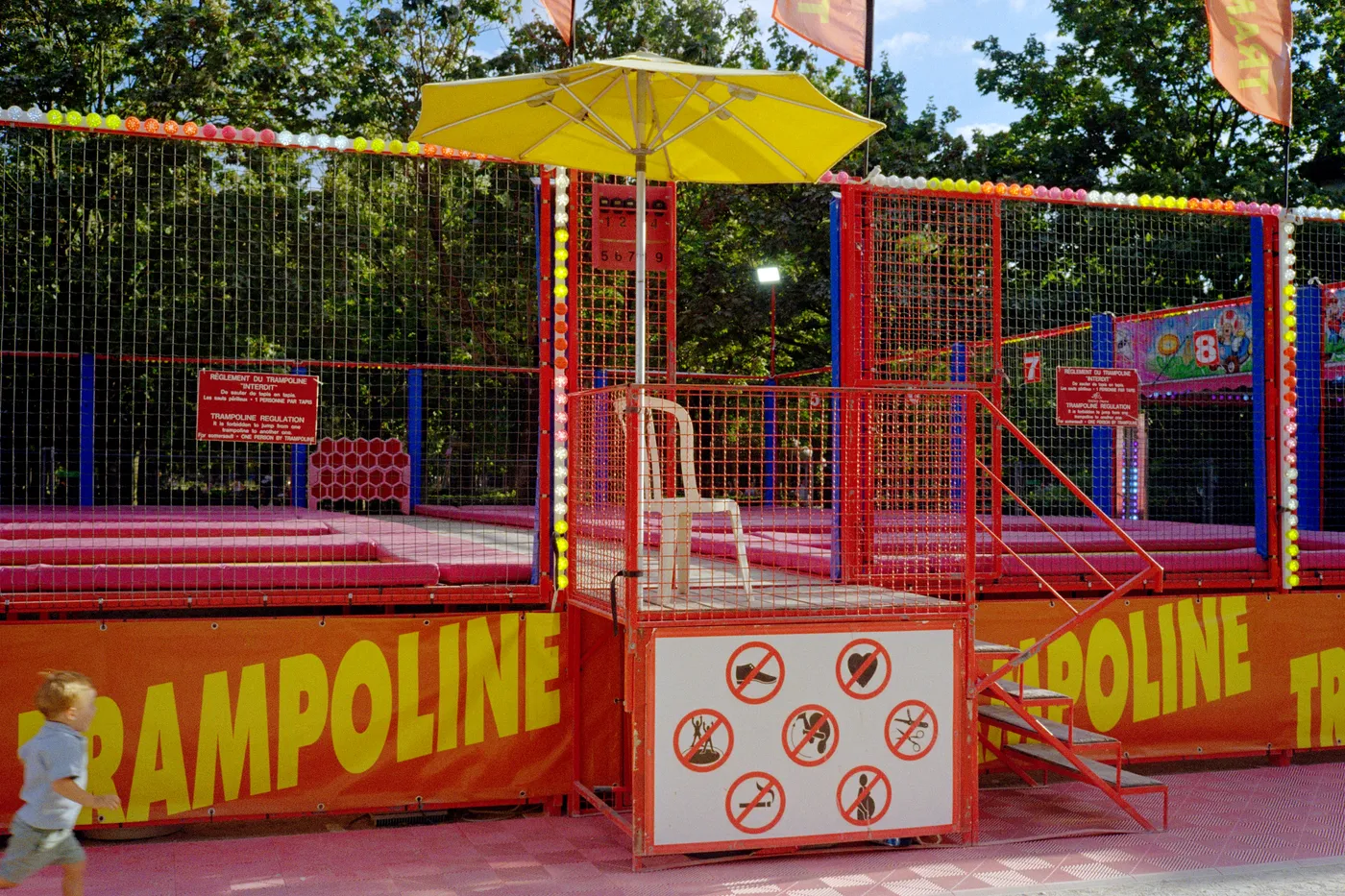 A fairground trampoline enclosure in flat daylight, orange banner against green trees.